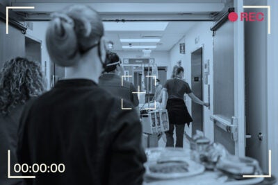 A group of medical professionals and workers wait for an elevator in a hospital hallway. Medical workers are wearing face masks and shields and have medical equipment and trays with them. The composition has a blue overlay and a recording frame to look like it’s being captured from a video camera.