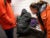 Two officials wearing bright orange winter jackets lean over to talk to an unhoused individual sitting on a bench in a Metro station. The individual is wearing an oversized black parka with the hood pulled up and rests on one arm. There are multiple bags on the bench near the person.