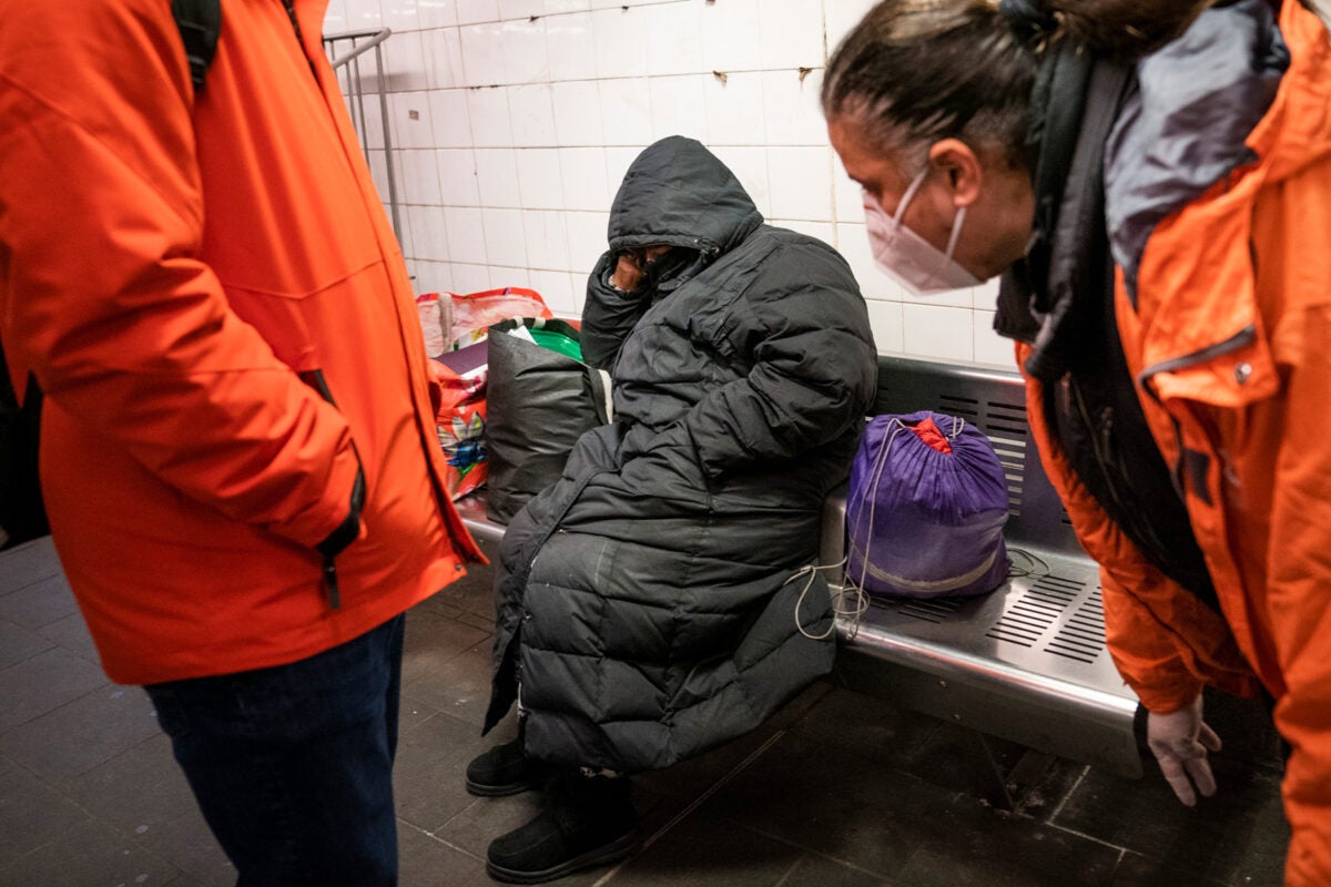 Two officials wearing bright orange winter jackets lean over to talk to an unhoused individual sitting on a bench in a Metro station. The individual is wearing an oversized black parka with the hood pulled up and rests on one arm. There are multiple bags on the bench near the person.