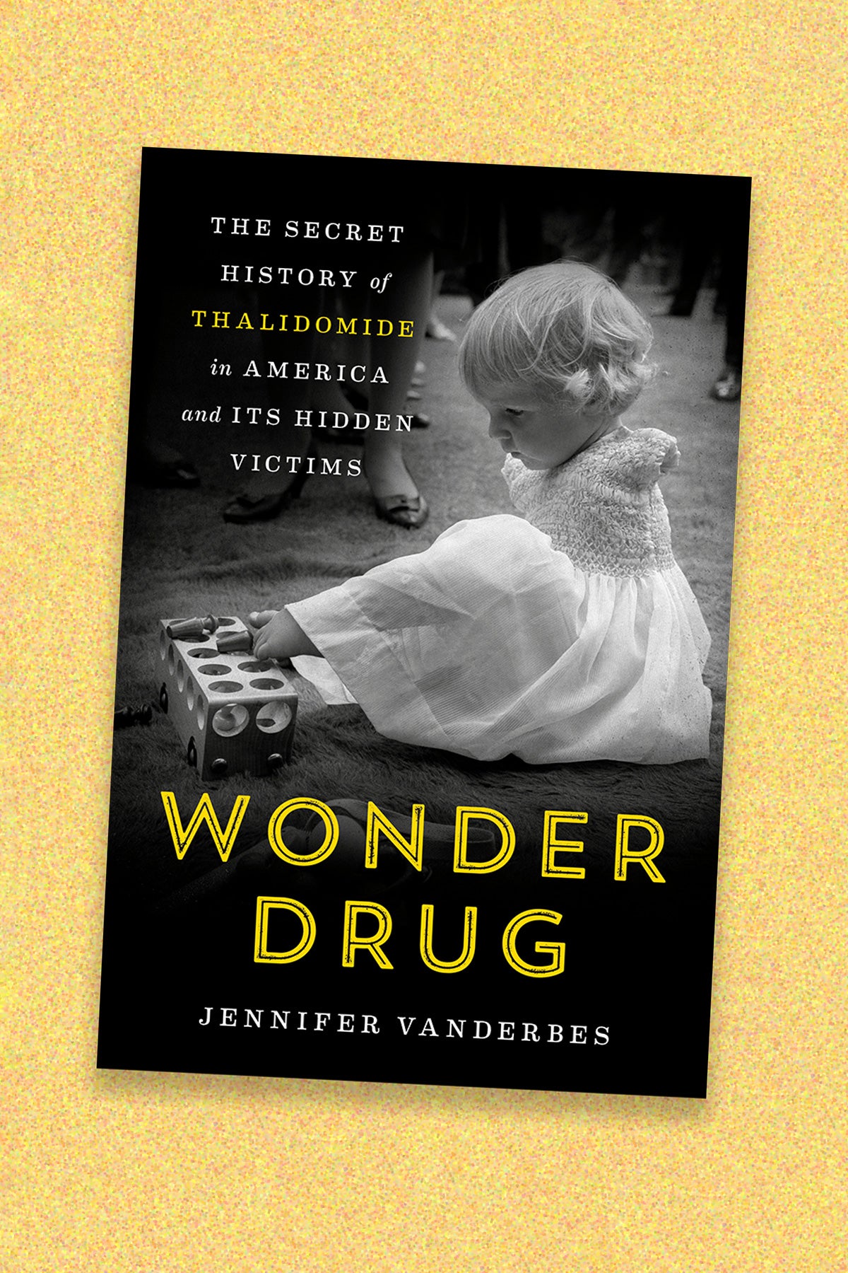 The book cover for “Wonder Drug: The secret history of thalidomide in American and its hidden victims” by Jennifer Vanderbes on a yellow speckled background. The book cover is black and has a black and white photo of a toddler-aged girl in a dress with no arms. She uses her toys to play with a wooden toy.