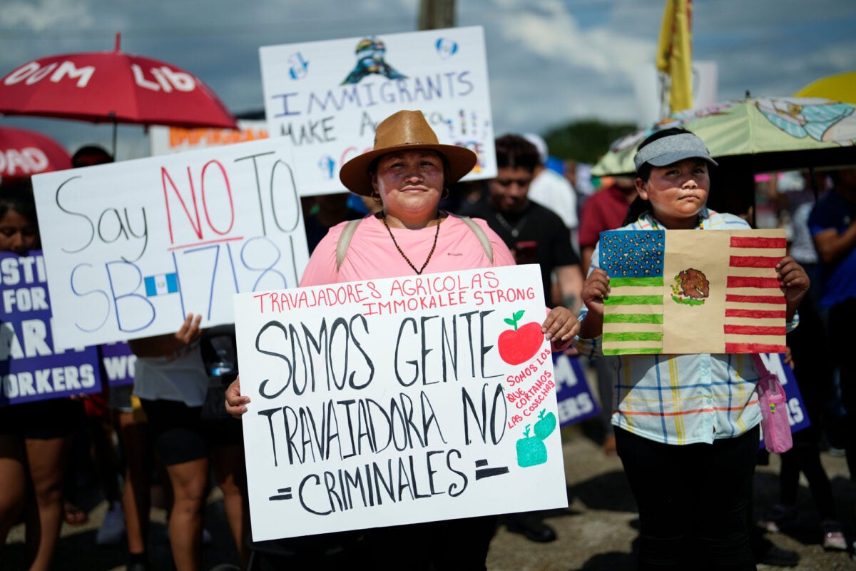 A female carries a sign that reads "We are working people, not criminals; we are the ones who harvest the crops; Immokalee farm workers strong” in Spanish at a large gathering of migrant farm and other workers. She wears a pink t-shirt and brown sun hat. On her right is a teenager holding a hand-colored flag that is half Mexico, half United States. On the main figure’s right is a sign that reads “Say no to SB 1718.”