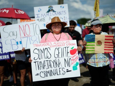 A female carries a sign that reads "We are working people, not criminals; we are the ones who harvest the crops; Immokalee farm workers strong” in Spanish at a large gathering of migrant farm and other workers. She wears a pink t-shirt and brown sun hat. On her right is a teenager holding a hand-colored flag that is half Mexico, half United States. On the main figure’s right is a sign that reads “Say no to SB 1718.”