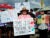 A female carries a sign that reads "We are working people, not criminals; we are the ones who harvest the crops; Immokalee farm workers strong” in Spanish at a large gathering of migrant farm and other workers. She wears a pink t-shirt and brown sun hat. On her right is a teenager holding a hand-colored flag that is half Mexico, half United States. On the main figure’s right is a sign that reads “Say no to SB 1718.”