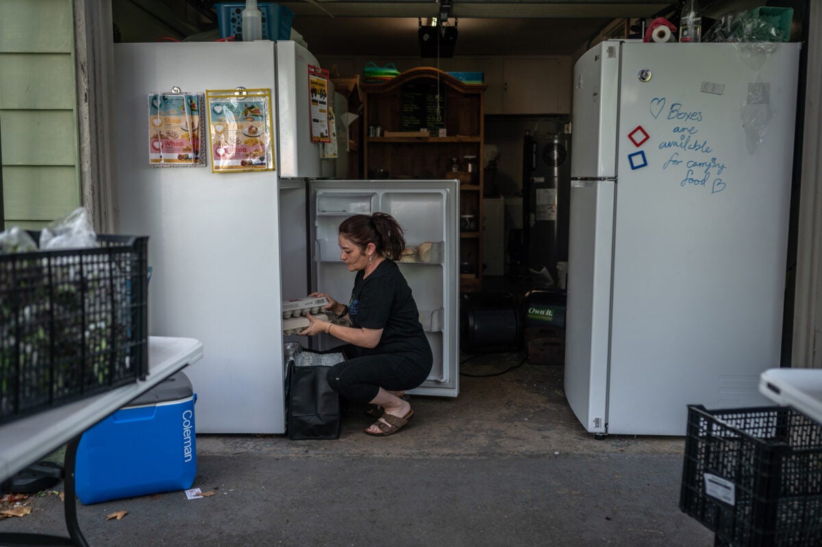 A woman squats in front of a fridge and holds two egg cartons in an open garage. She has a black freezer bag in the foreground, wears black clothes, and brown sandals. There is another fridge, folding tables with black crates and a blue cooler.