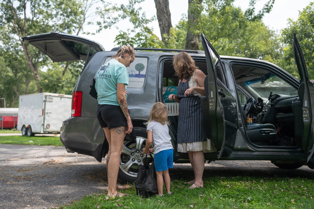 A young woman, a four year old girl and a middle aged blonde woman stand in front a black SUV with its doors open. The car is loaded with fresh fruit and other food being delivered.