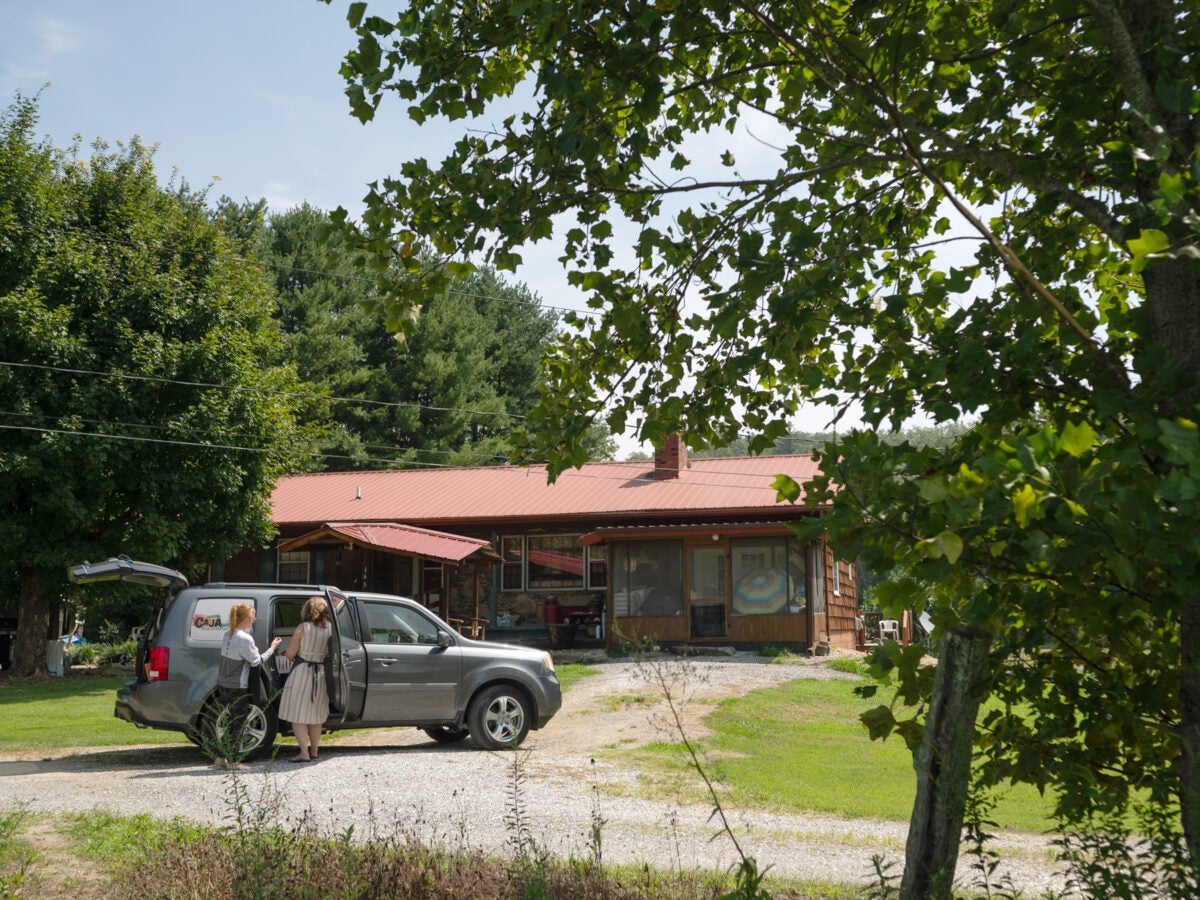 Two women stand near in front of an open side door of a black SUV. The trunk of the car is also open. The logo for “Caja Solidaria” is applied on the rear window. The car is parked in a dirt driveway in front of a stone ranch house with a red tin roof in rural North Carolina. The front sun room of the house has two open umbrellas, plastic bins and other stuff. A tree with bright green leaves is in the front right of the image.