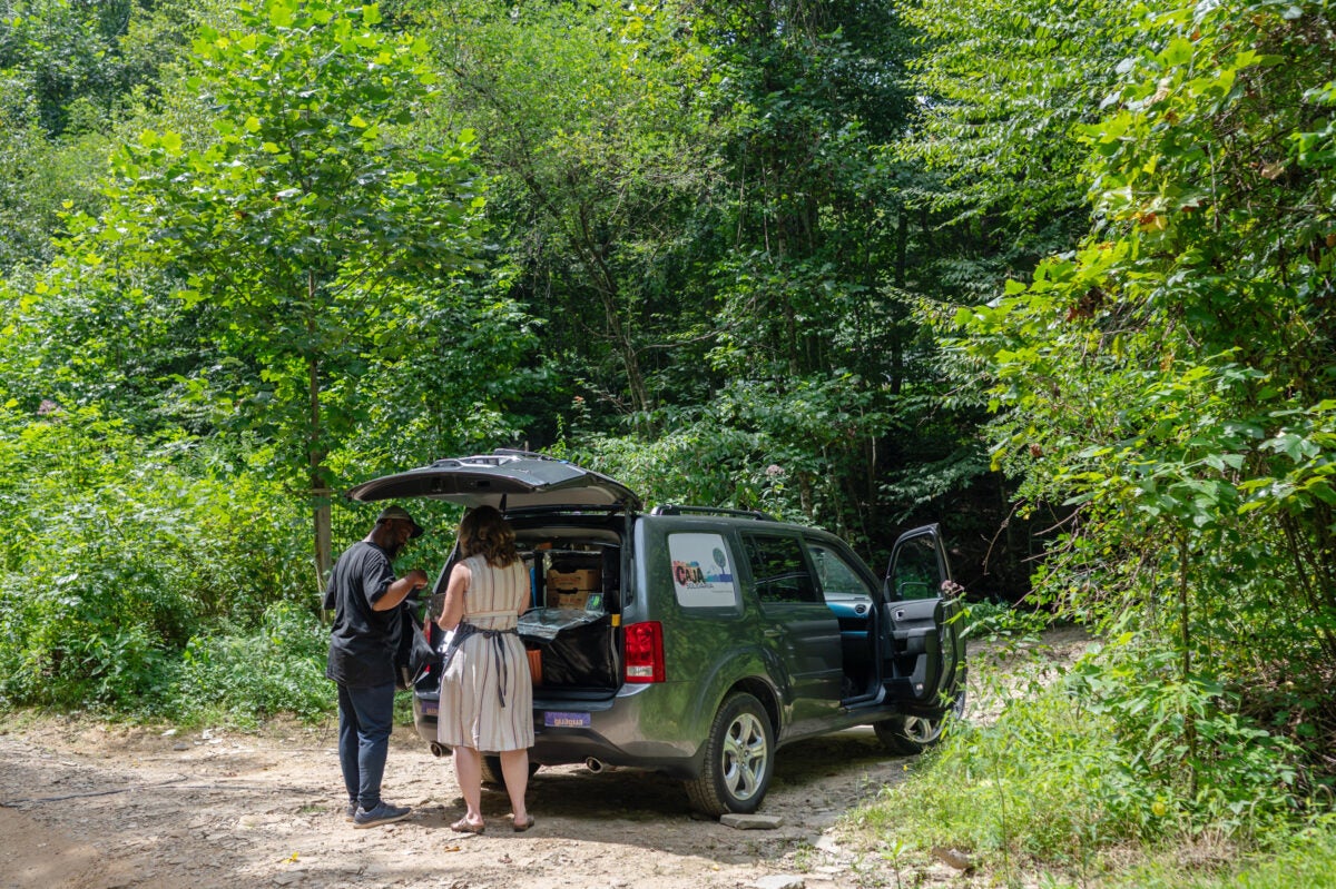 A black man and white woman stand on a dirt road in rural North Carolina in front of an open SUV trunk filled with fresh fruit and other food.