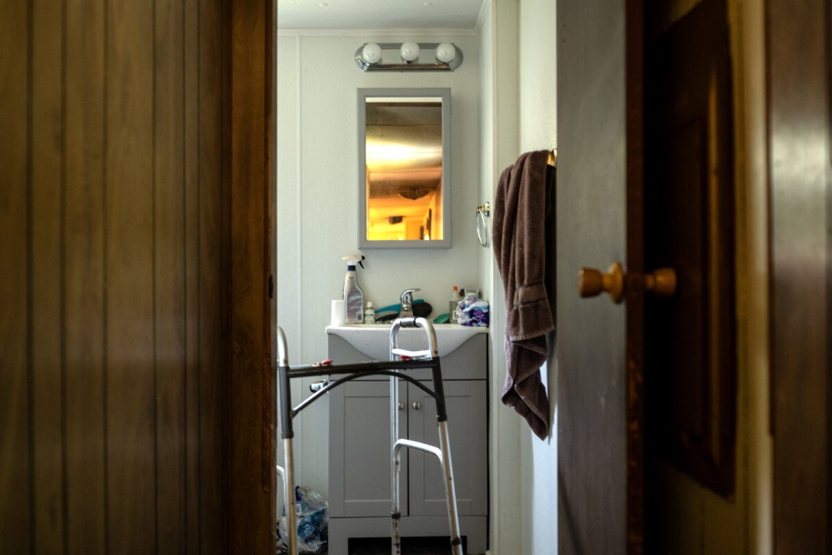 A walker sits in the doorway of a recently renovated bathroom with a new grey vanity, white mirror and lights. The sink is covered with standard bathroom items: toilet paper, contact solution, cleaner, etc. A brown towel hangs from a towel rod on the right.