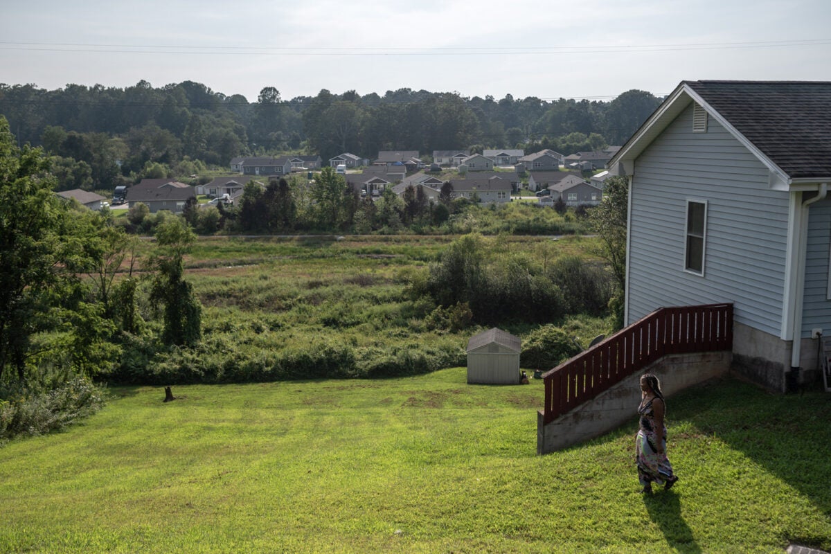 A Black woman walks in her large mowed grass lawn in rural North Carolina. Other houses in a far off subdivision are visible in the background.