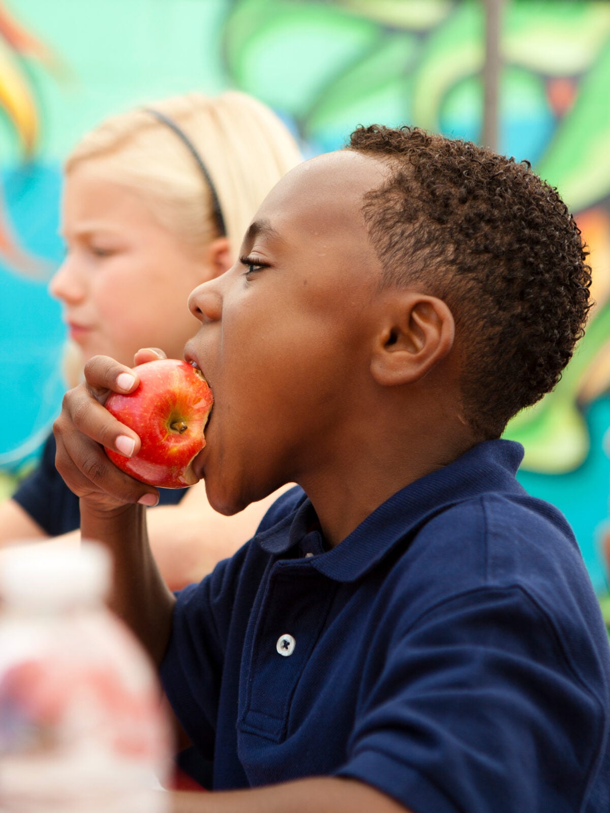 Two elementary-aged children eat apples in a school cafeteria. They are wearing blue-polo shirts and there is a bright mural in the background.