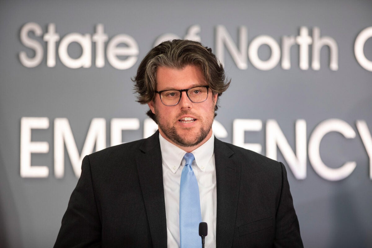 Kody Kinsley, North Carolina Health and Human Services Secretary, speaks into a microphone at a press conference. He is wearing a dark suit with a light blue tie and glasses.