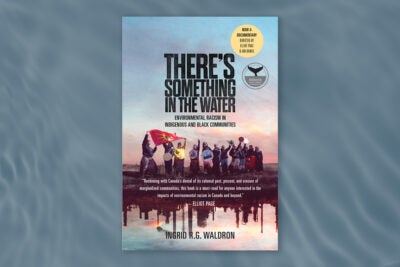 Book cover for “There’s something in the water: Environmental racism in indigenous and black communities” by Ingrid R.G. Waldron. The cover is composed on a photo of a group of indigenous people with drums and flags raised, standing near a body of water. Reflected in the water are industrial facilities and steam stacks. The book cover has two stickers in the top right: “no a documentary directed by Elliot page and Ian Daniel” and “Atlantic Book Awards Winner.” The book cover itself is on a dark watery background image.