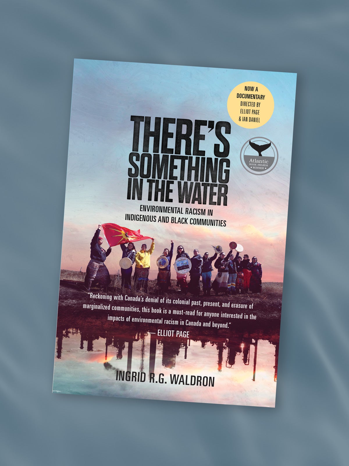Book cover for “There’s something in the water: Environmental racism in indigenous and black communities” by Ingrid R.G. Waldron. The cover is composed on a photo of a group of indigenous people with drums and flags raised, standing near a body of water. Reflected in the water are industrial facilities and steam stacks. The book cover has two stickers in the top right: “no a documentary directed by Elliot page and Ian Daniel” and “Atlantic Book Awards Winner.” The book cover itself is on a dark watery background image.