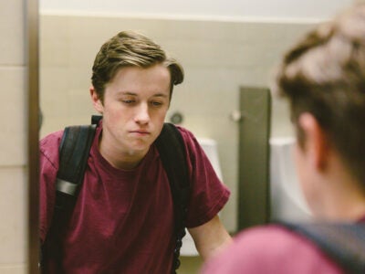A depressed-looking, white teenage boy wearing a maroon shirt and black backpack looks at himself in a school bathroom mirror.