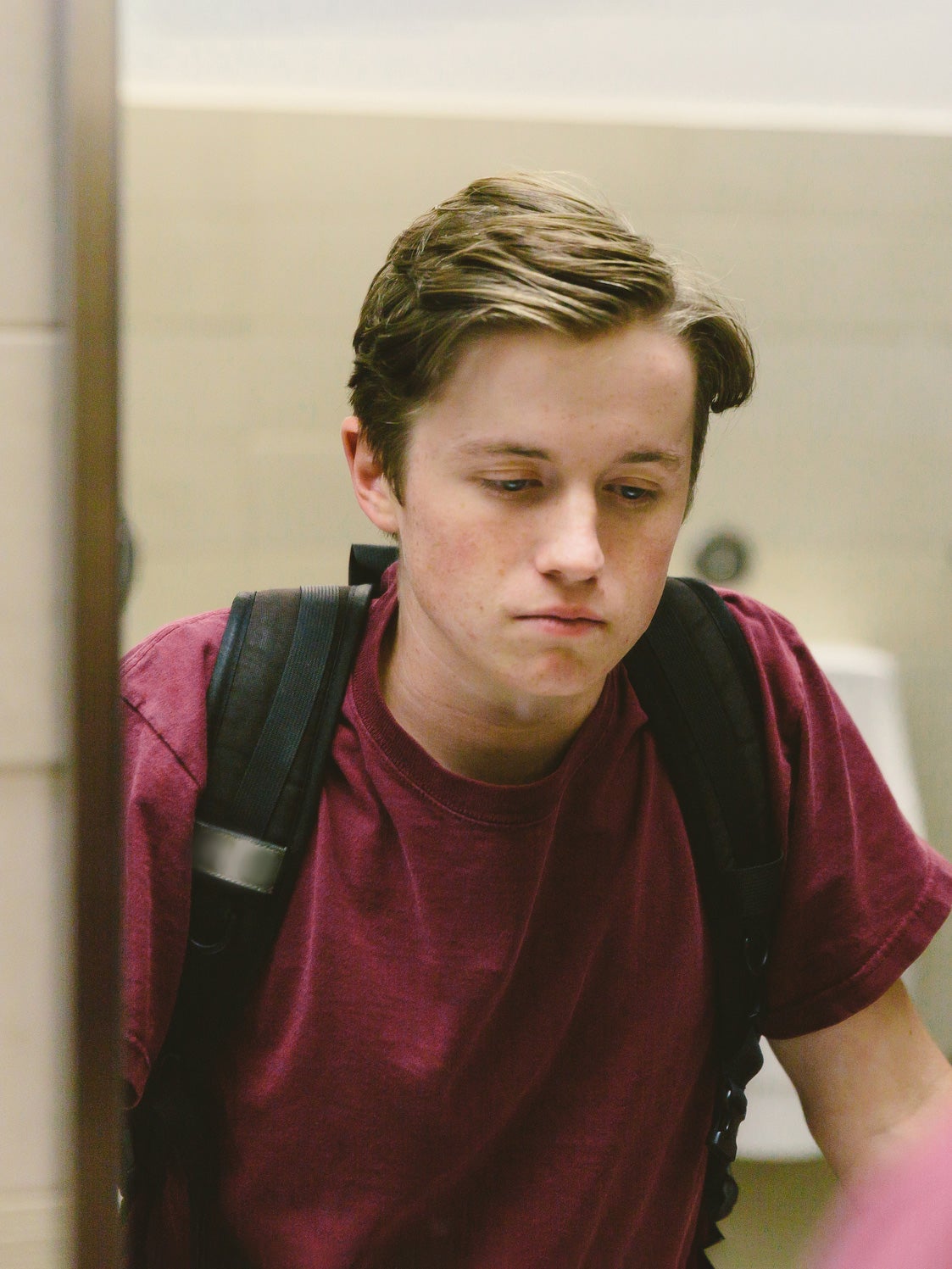 A depressed-looking, white teenage boy wearing a maroon shirt and black backpack looks at himself in a school bathroom mirror.
