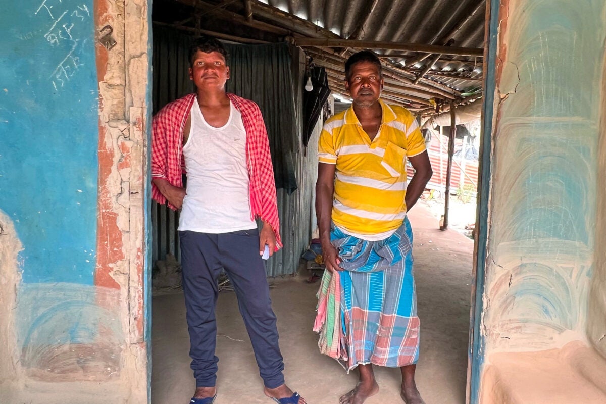 Two middle-aged Indian brothers stand in a doorway in rural India. One wears a red shawl, white tank top, blue joggers and sliders. The other wears a yellow and white striped polo shirt and a blue, red, and grey patterned skirt.