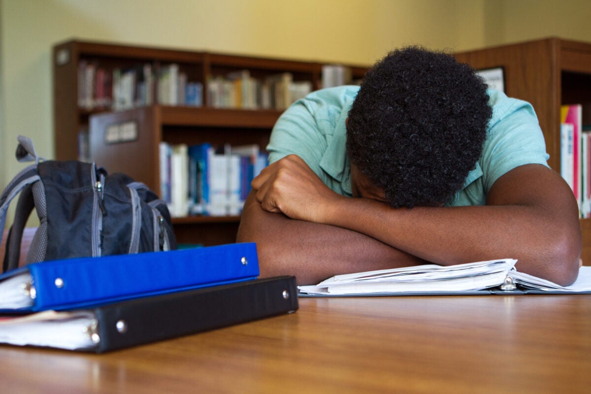 A teenager rests his head in his forearms and sleeps at a school library table. Three binders are on the table and his book bag sits in a chair.