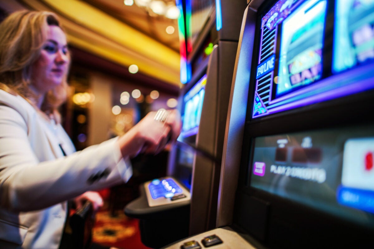 A blonde woman plays the slot machines at a casino. The blue glow of the slot machine screen next to her shines in the foreground.