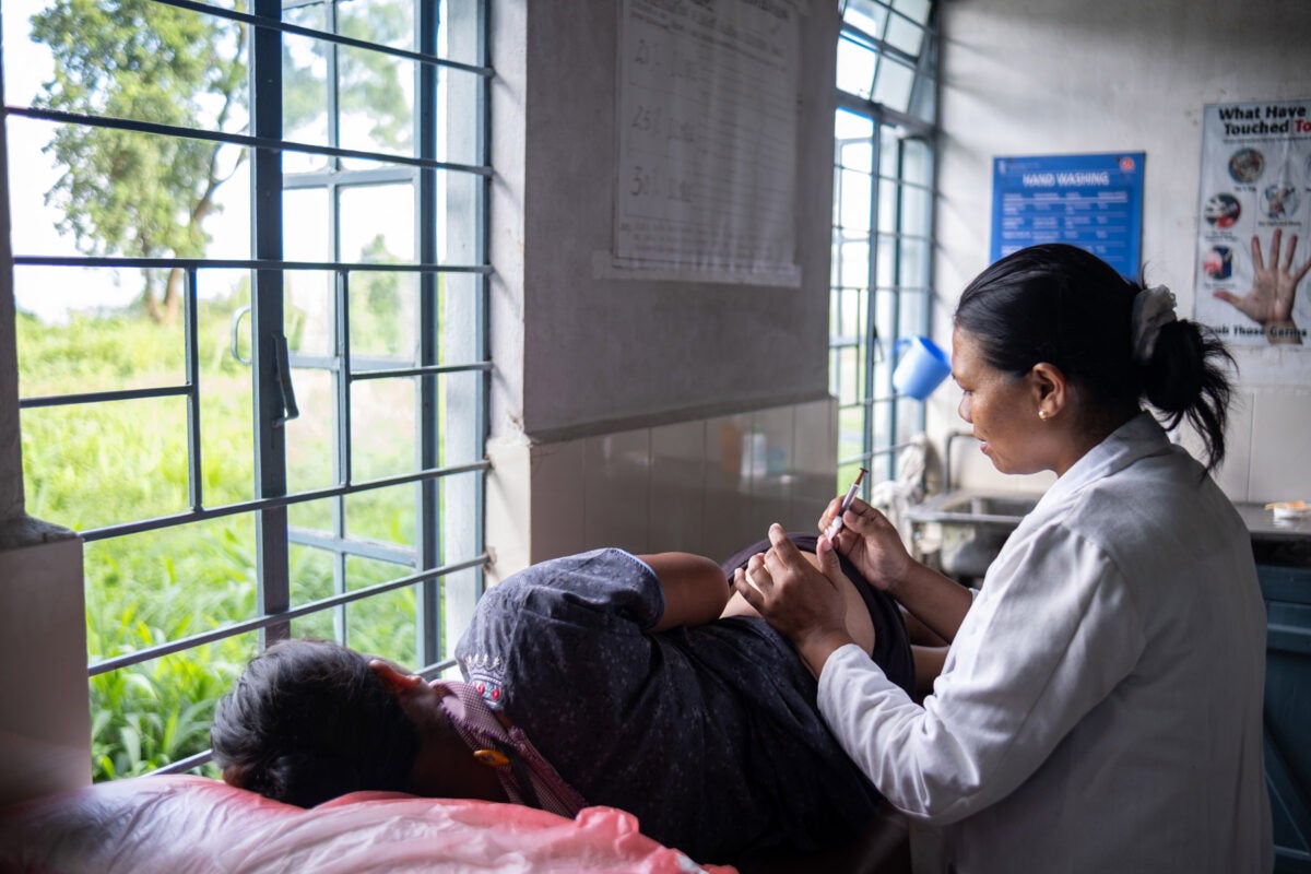A female midwife wearing a white coat inserts a vaccine vial into the hip of a female patient who is resting on her side, looking out a window into a green field, at a health clinic in northeastern India.