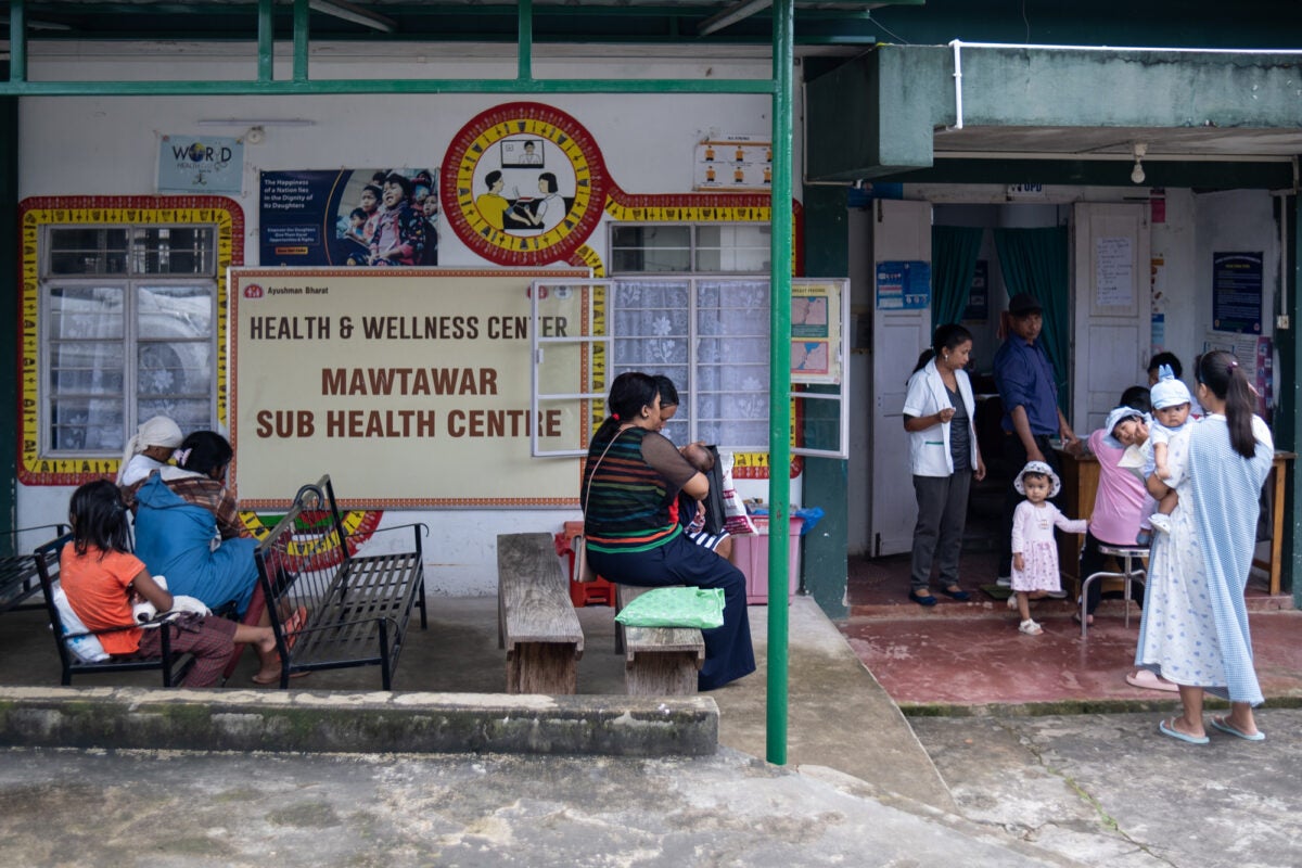 The exterior of Mawtawar Sub Health Centre in northeastern India. Mothers with children sit on benches on a concrete patio and others stand in a reception line. The building has two windows with curtains, a large sign and blue and red graphics around the windows.