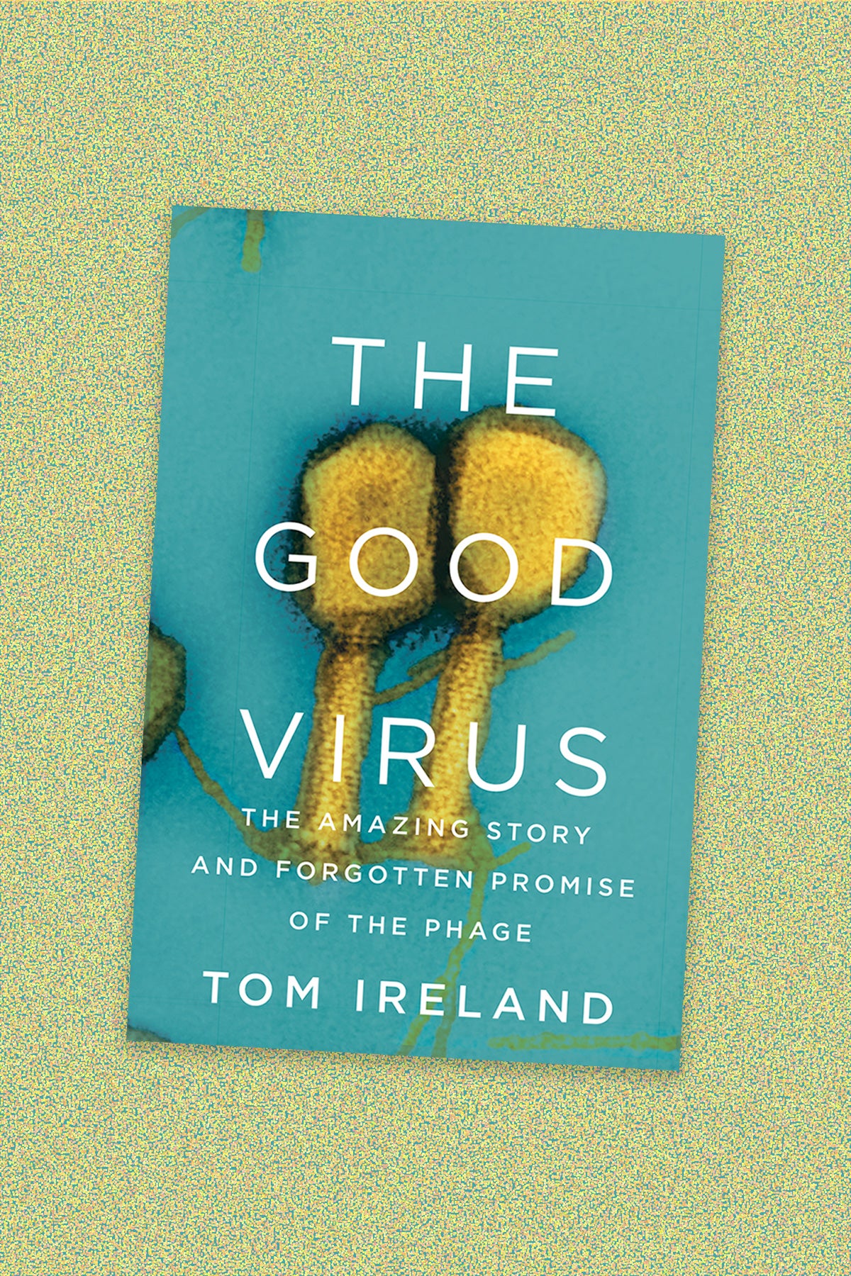Book cover of “The Good Virus: The amazing story and forgotten promise of the phage” by Tom Ireland. The book cover shows a microscopic image of a yellow phage virus on a teal background. The cover text is white all-caps. The book cover sits on a yellow and teal-speckled background.