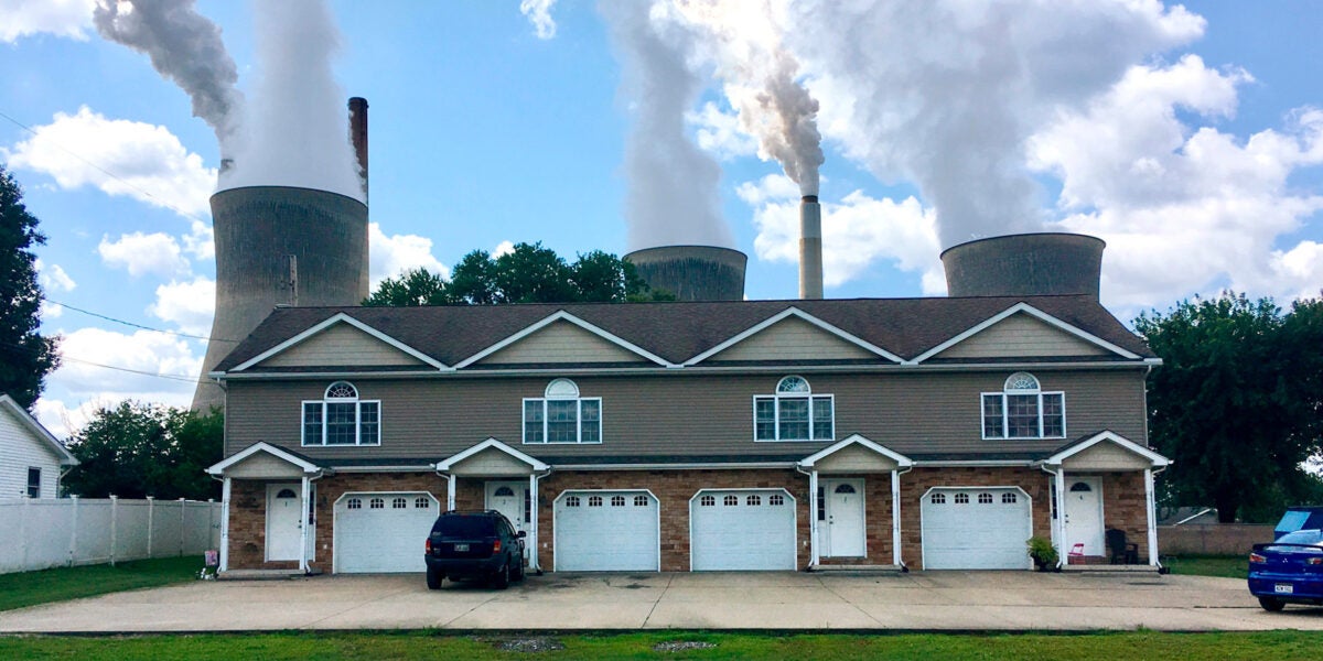 A row of townhomes in West Virginia with coal and nuclear power plants immediately behind them. They steam stacks blow air into the bright blue sky.
