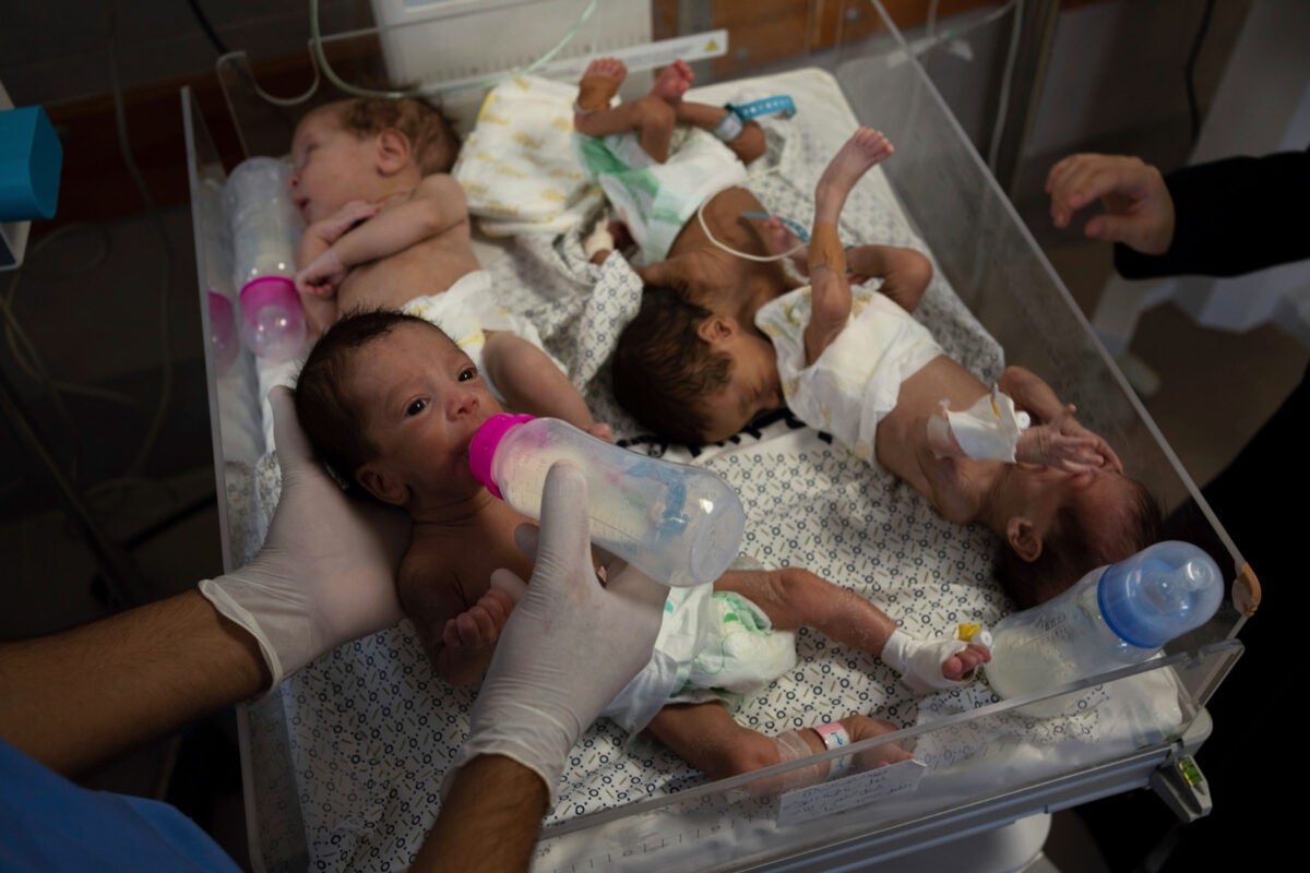 Four premature babies share one plastic hospital bassinet Shifa Hospital in Gaza city. The infant in the bottom left is being fed by a gloved medical worker out of a bottle with a pink top. The bassinet has a patterned sheet, and diapers and bottles are laid among the infants.