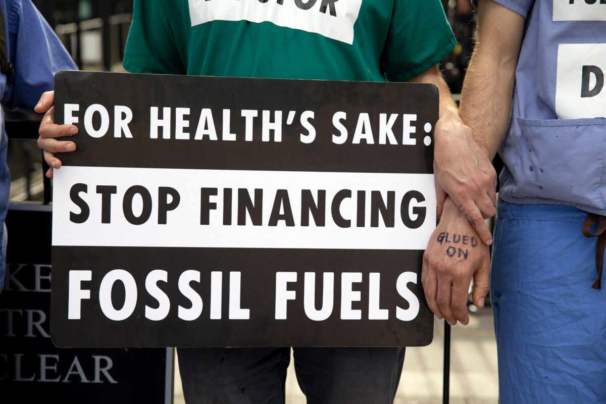 Healthcare professionals stand in protest. One holds a black and white sign that reads “For Health’s Sake: Stop Financing Fossil Fuels.”
