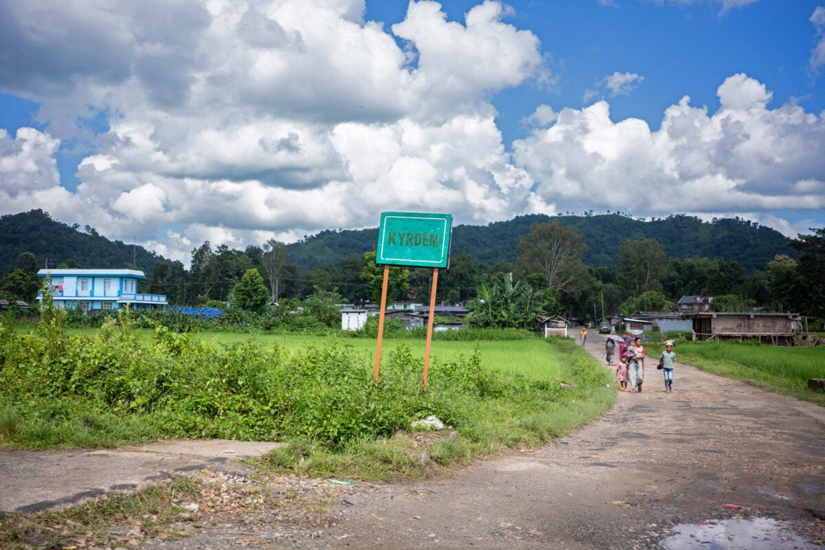 A green road sign reads "Kydrem" on a dirt road along a field in rural northeastern India. A family walks the path in the distance.