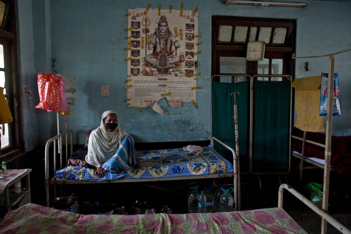 A female tuberculous patient sits on a bed at a TB hospital in Gauhati India. She wears a mask. The room has patterned linens and green hospital curtains.