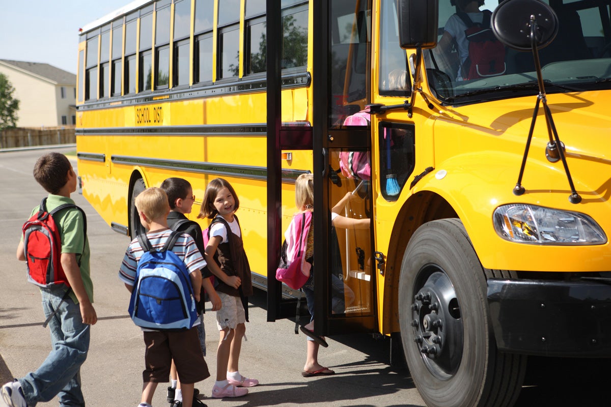 Young children stand in a line, waiting to get onto a yellow school bus.