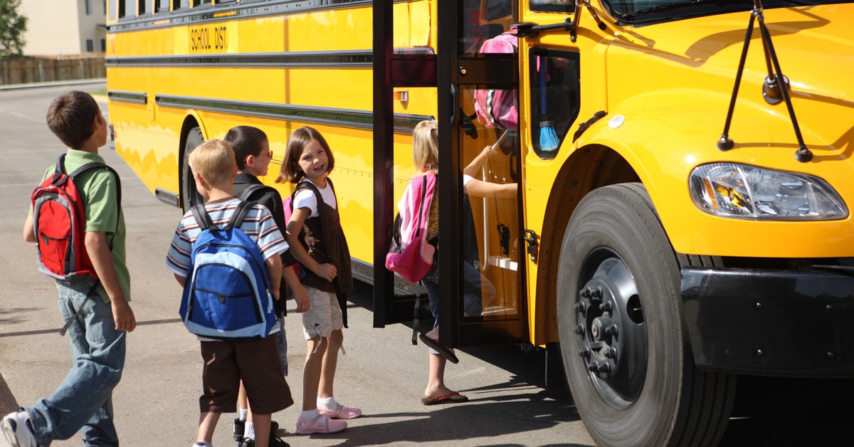 Young children stand in a line, waiting to get onto a yellow school bus.