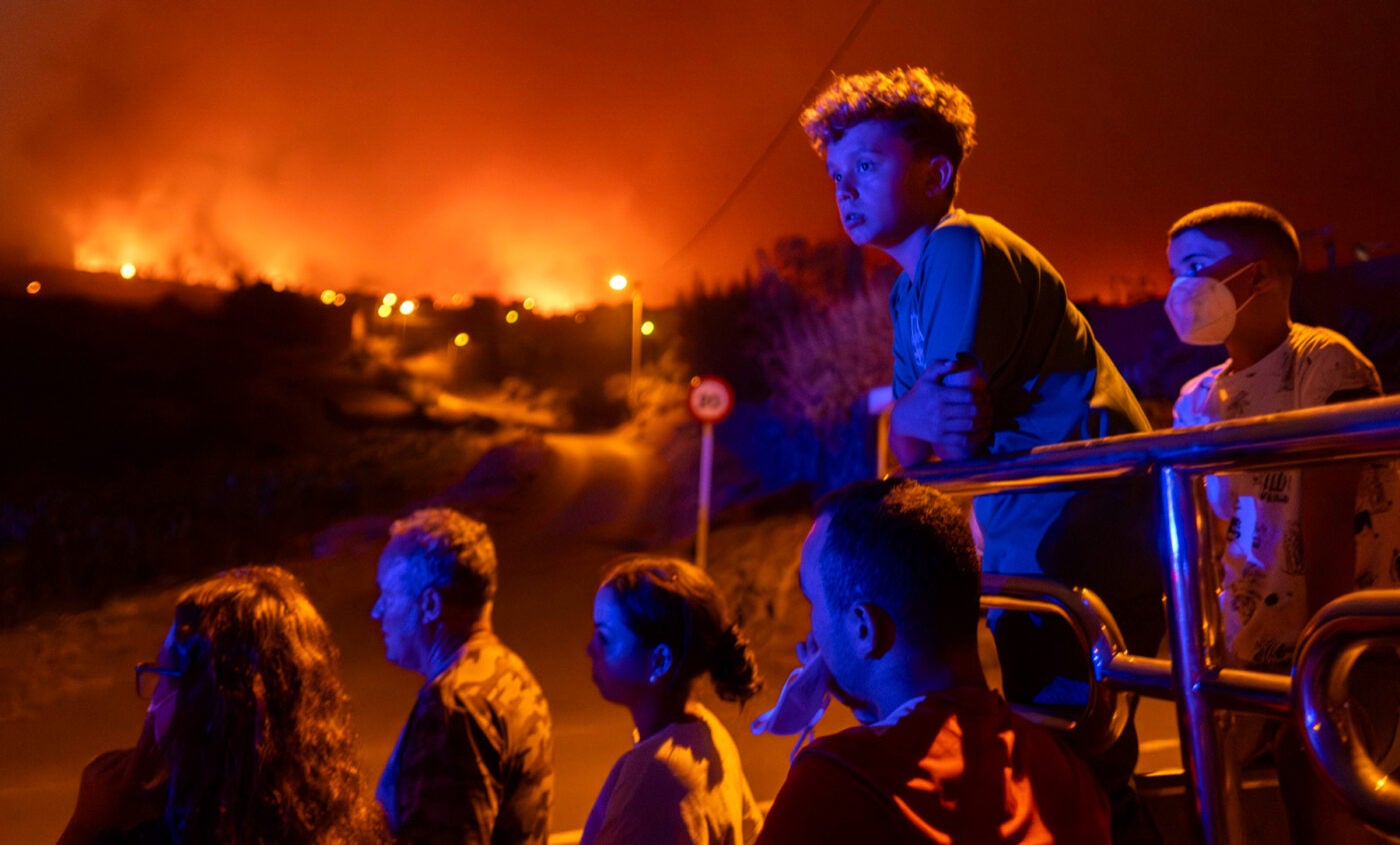 Two boys and other family members stand in the bed of a vehicle, watching wildfires in the distance in their neighborhood of Benito’s village, Spain. The sky glows orange and a blue glow is cast on the figures’ faces.