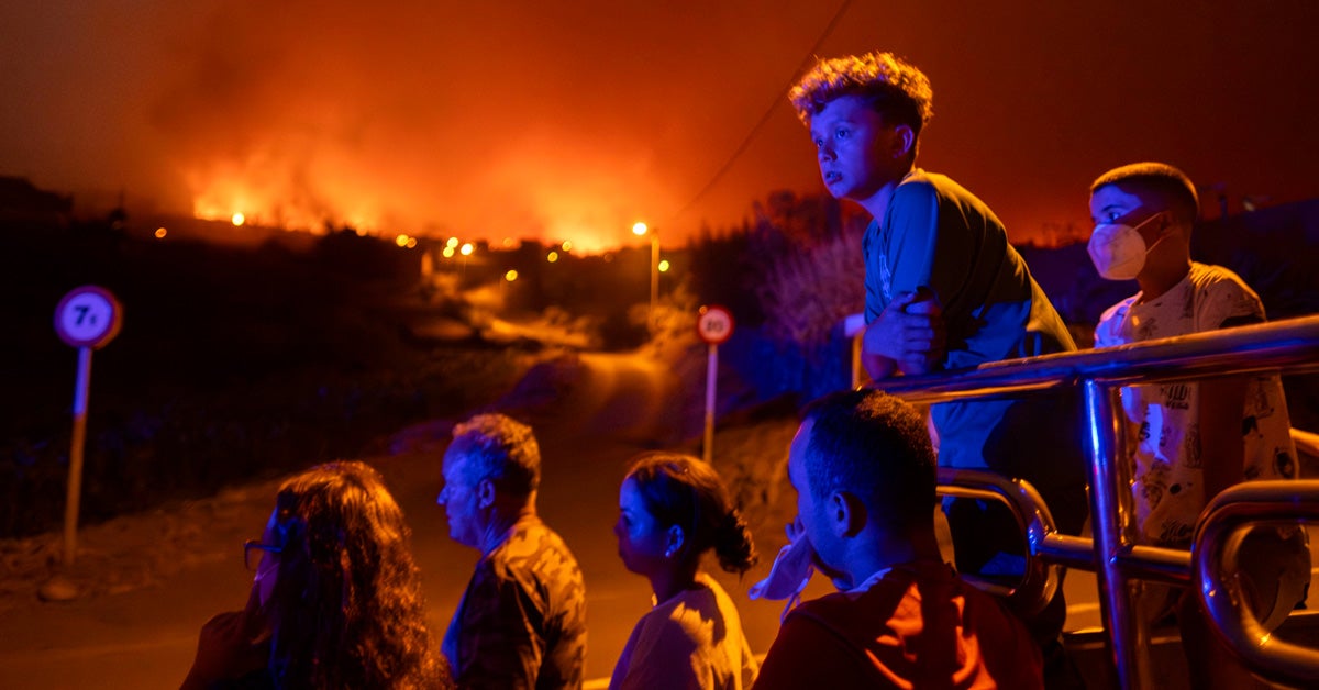 Two boys and other family members stand in the bed of a vehicle, watching wildfires in the distance in their neighborhood of Benito’s village, Spain. The sky glows orange and a blue glow is cast on the figures’ faces.
