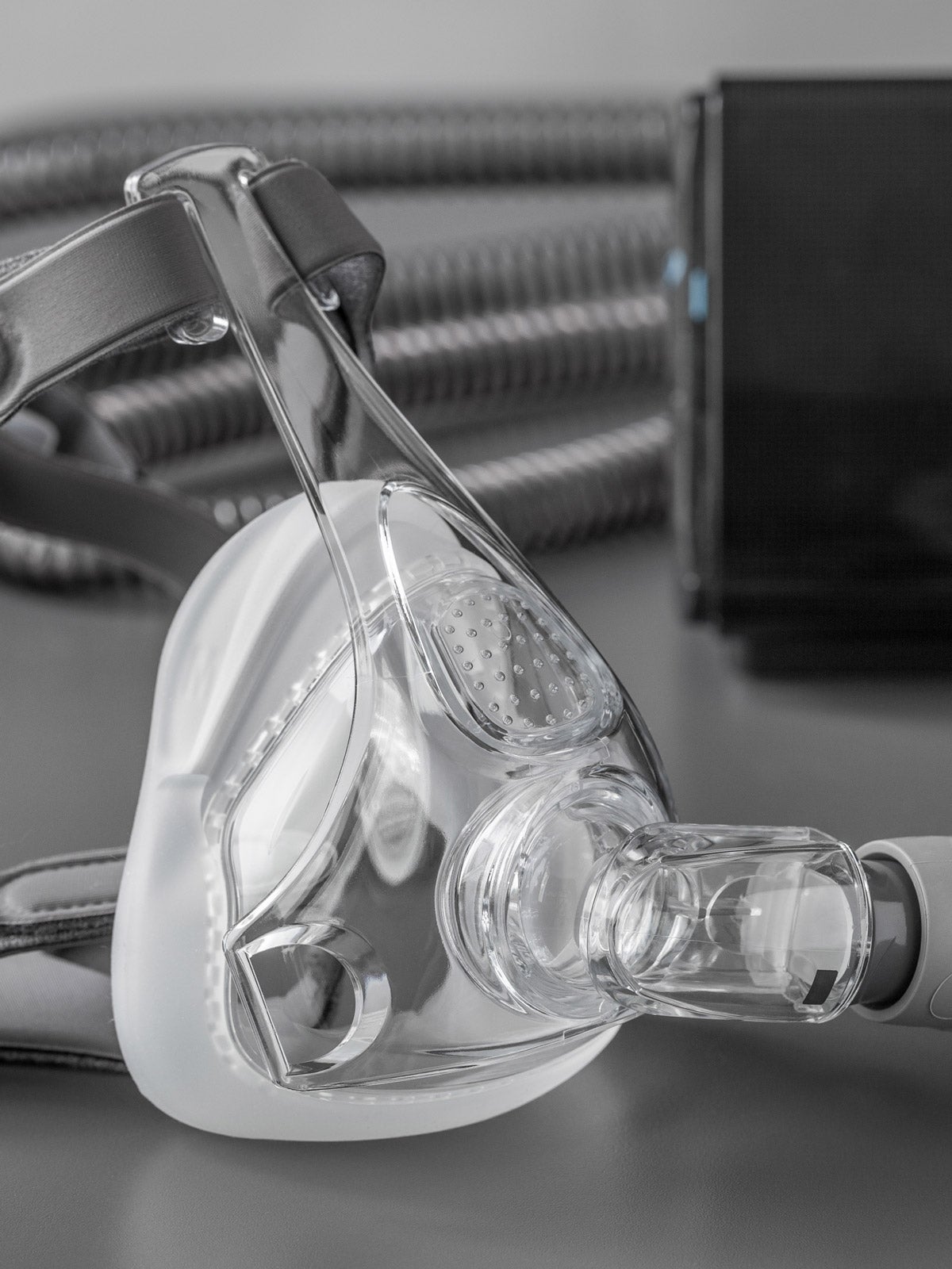 Clear sleep apnea CPAP mask with grey straps and tubes on a grey table. The black CPAP machine is in the background.