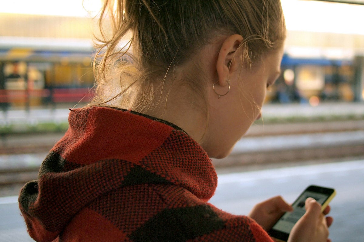 A female teen in a red plaid hoodie interacts with her phone screen at a train station.