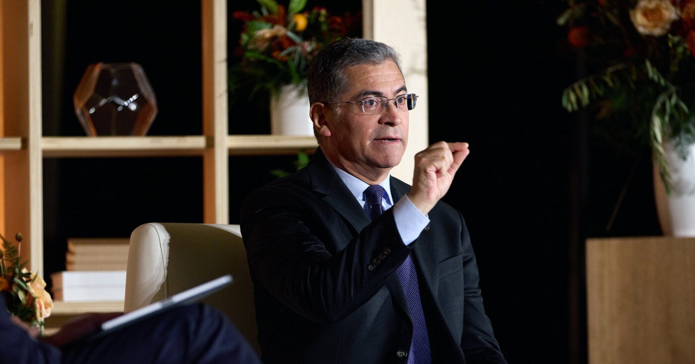 Secretary of Health and Human Services Xavier Becerra speaks at a media conference. He is seated in a cream arm chair and speaks to the audience. He wears a dark blue suit, tie and thin-rimmed glasses.