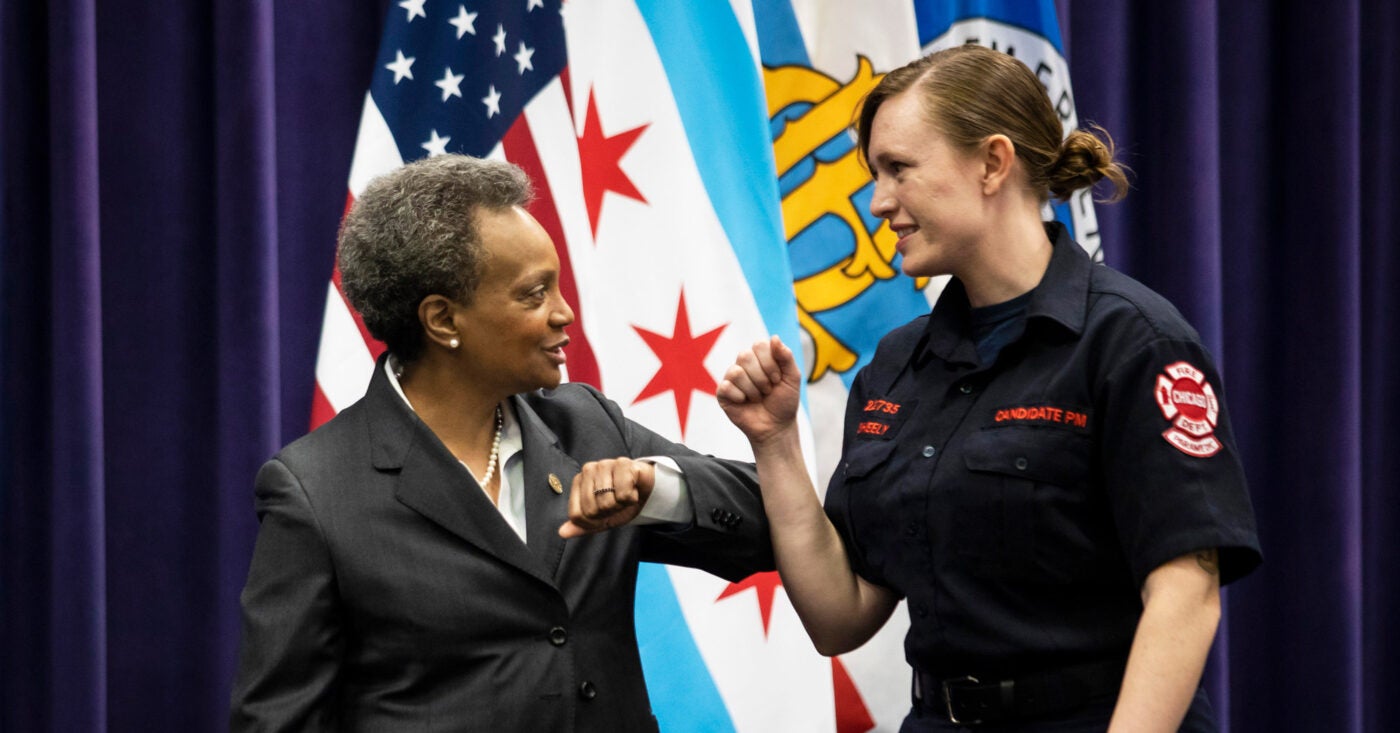 A female firefighter in dress fatigues bumps elbows with Mayor Lori Lightfoot at her graduation ceremony. The two stand in front of an American flag, a city of Chicago flag and two other flags.