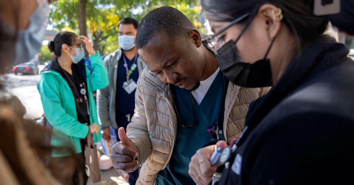 A black immigrant works with a health care worker to fallout a form on a clipboard in an outdoor urban area. Other health care workers stand in the background.