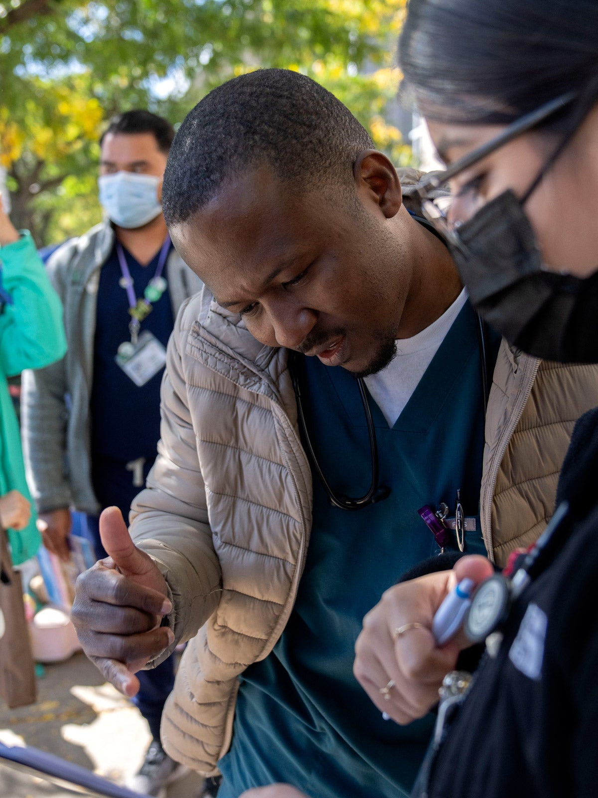 A black immigrant works with a health care worker to fallout a form on a clipboard in an outdoor urban area. Other health care workers stand in the background.