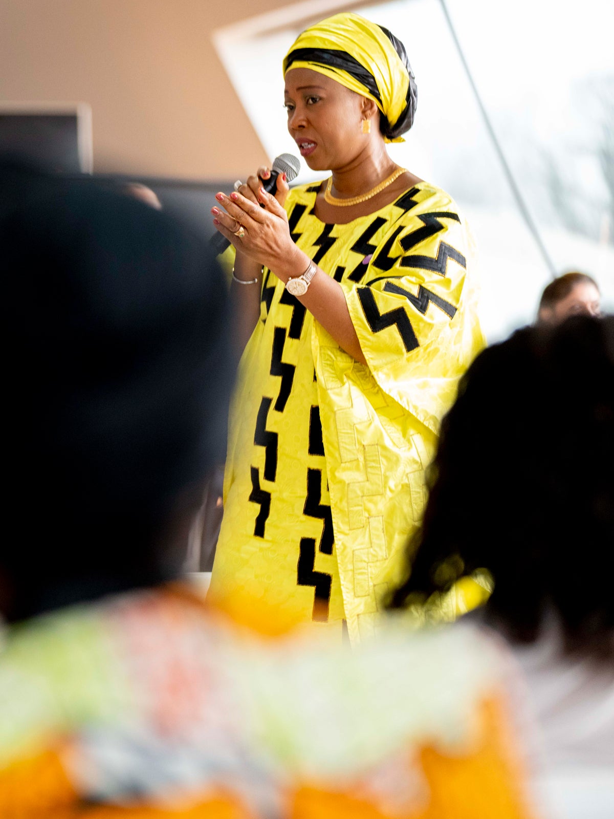 Fatima Maada Bio speaks into a microphone to a group of people. She is wearing a robe and headdress that are both bright yellow with black highlights.