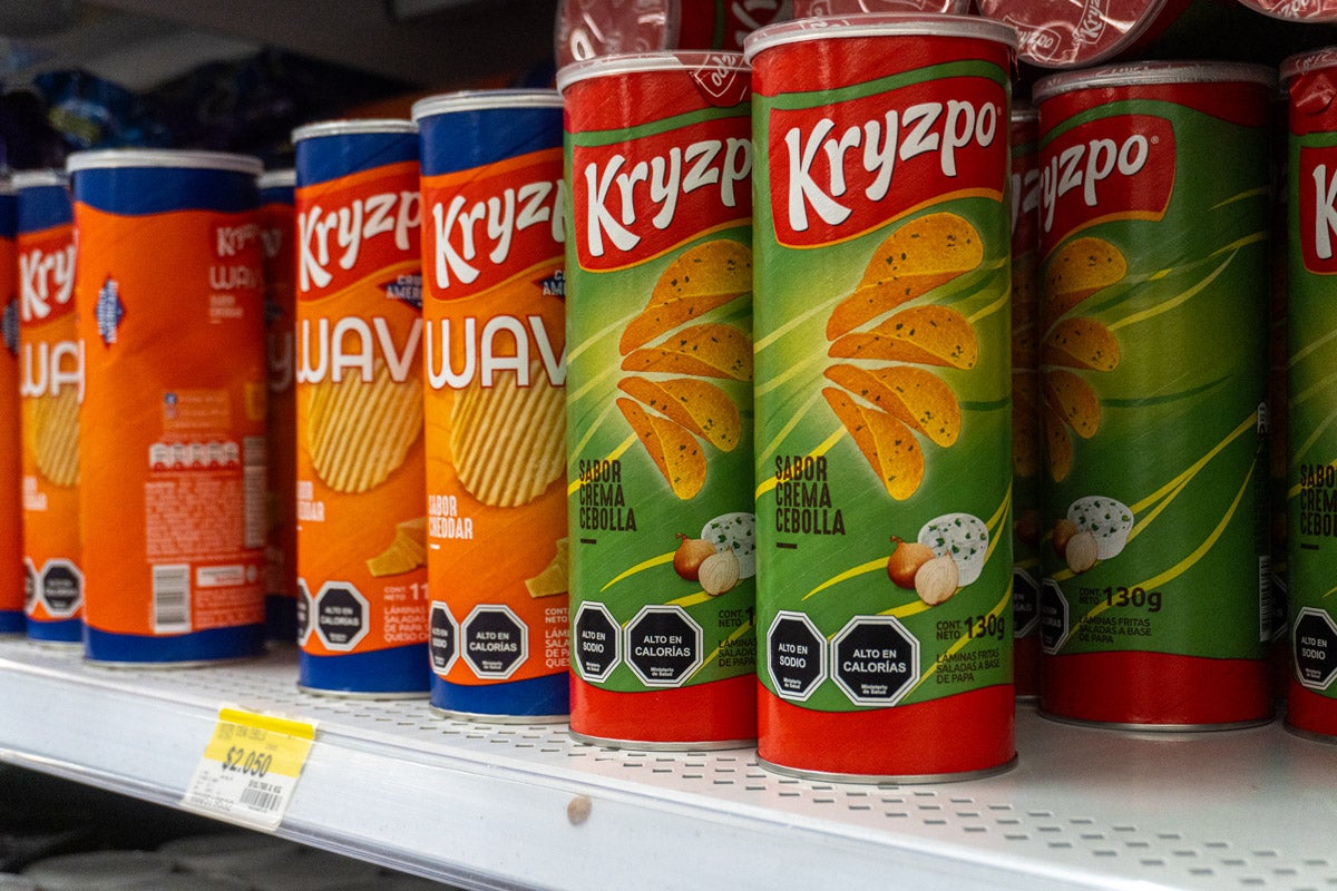 Cans of Kryzpo brand potato chips, labeled in Spanish, line a supermarket shelf. Black labels on each can warn they are "high in sodium" and "high in calories."