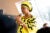 Fatima Maada Bio speaks into a microphone to a group of people. She is wearing a robe and headdress that are both bright yellow with black highlights.