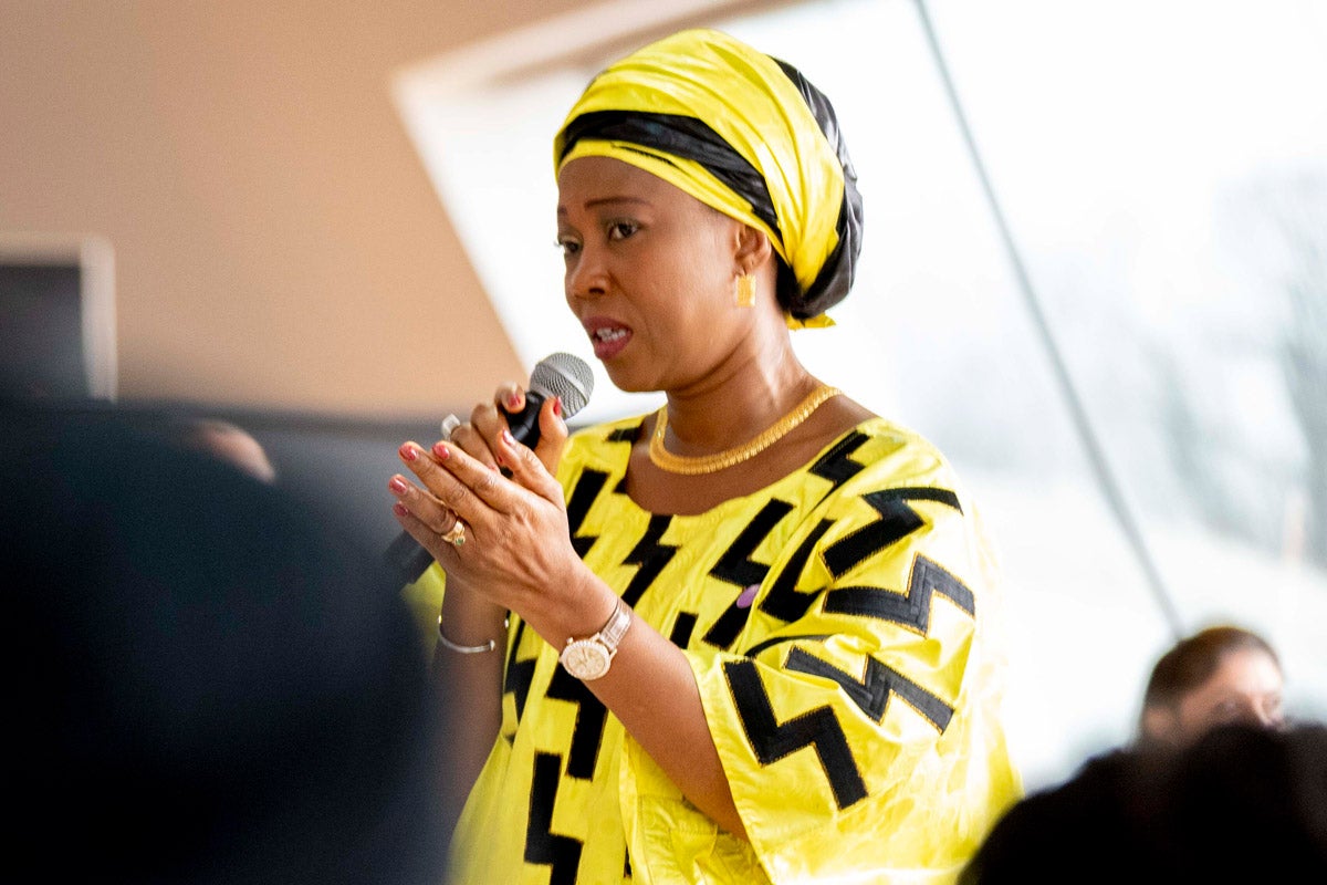 Fatima Maada Bio speaks into a microphone to a group of people. She is wearing a robe and headdress that are both bright yellow with black highlights.
