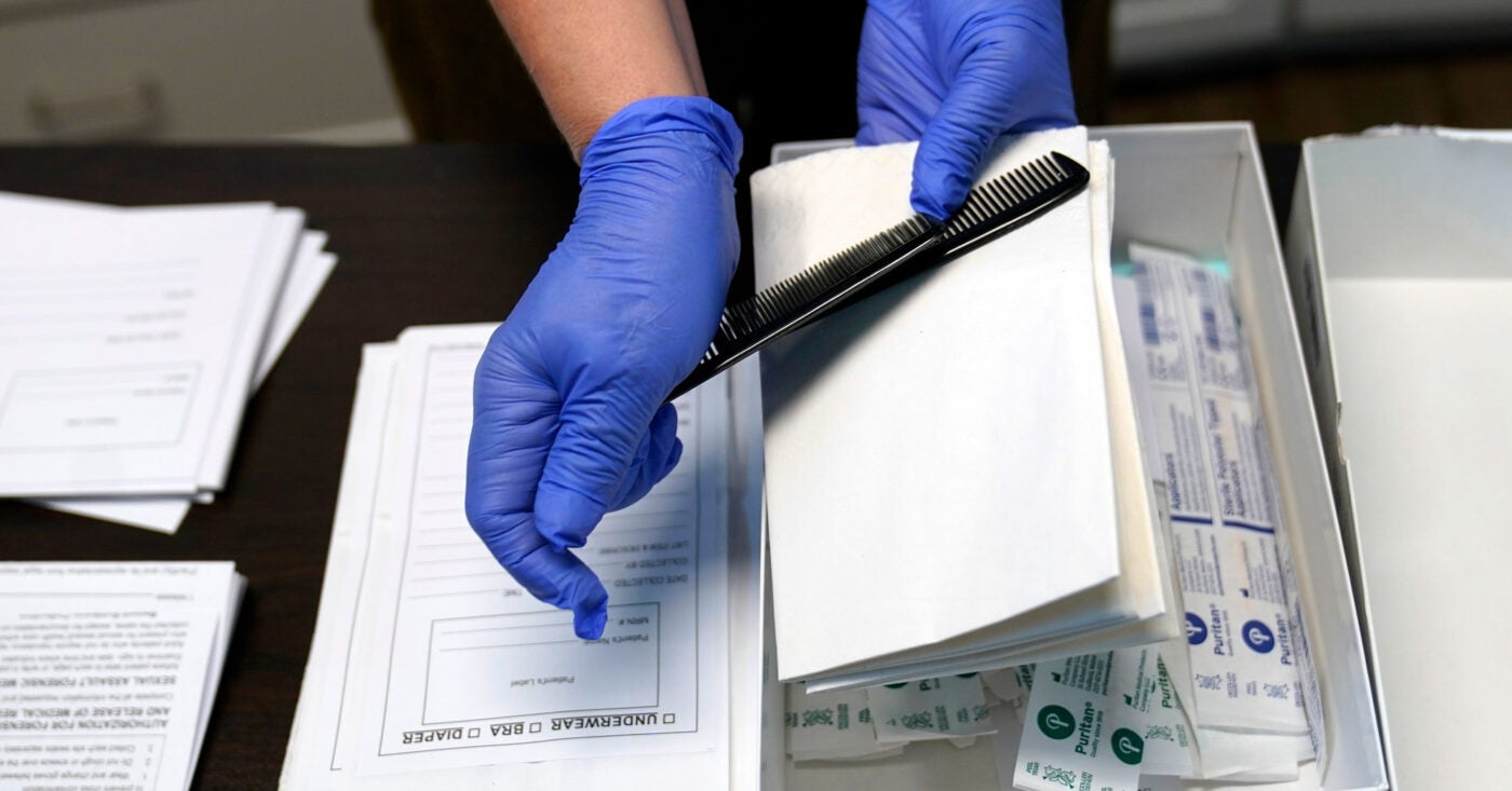 Two latex gloved hands hold a black comb as part of a displayed sexual assault evidence kit (rape kit). On the table are a bunch of swabs in a box, small envelopes and labels for clothes.