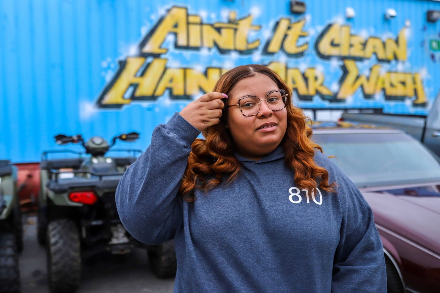Dionna Brown stands outside a car wash, staring at the camera while placing some hair behind her ear. The words "Ain't it clean hand car wash" in yellow block type on a bright blue wall are behind her.