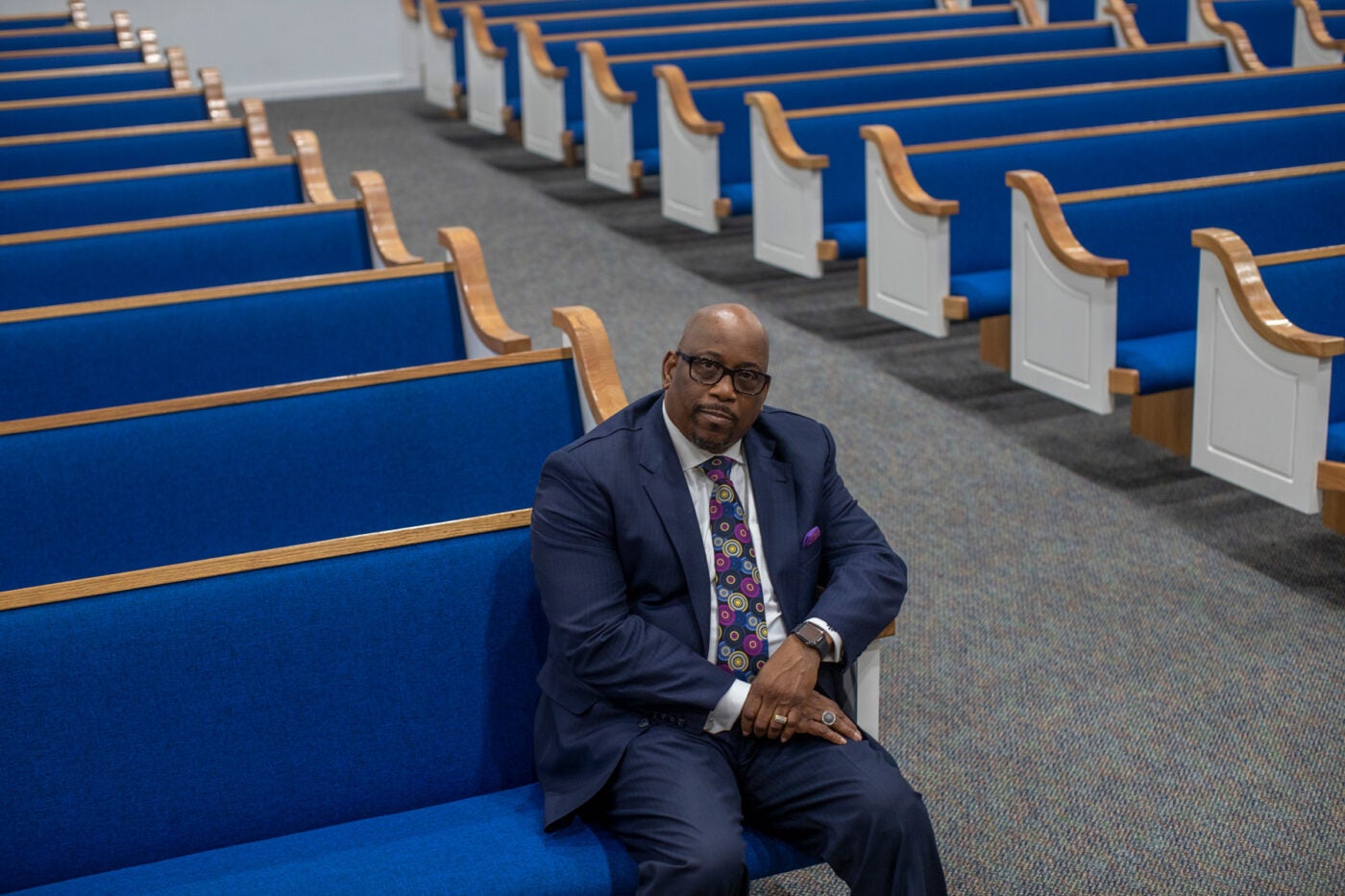 Pastor Allen Overton sits in a wooden pew with bright blue upholstery in his church. The pews cascade behind him in two rows.