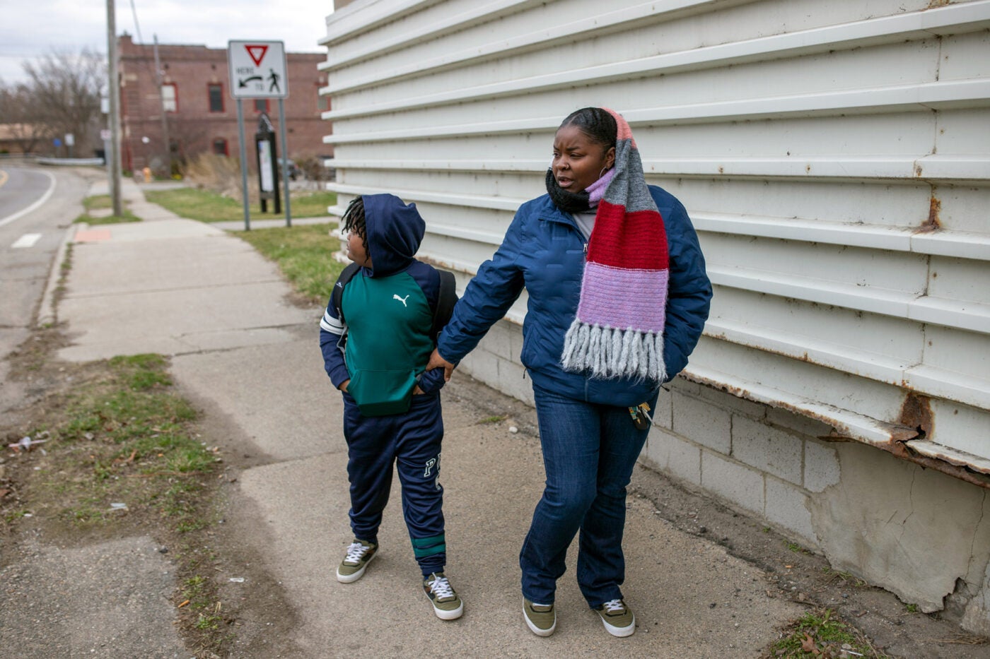 A woman walks with her son on a sidewalk by a white sided building in Flint, Michigan. She wears a blue coat and brightly colored knit scarf. He wears puma sweats with his hood up.