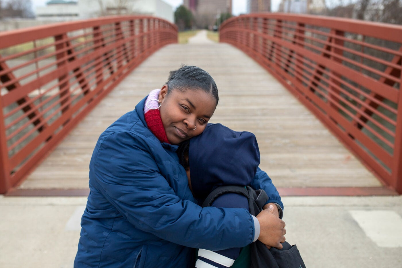 Shaketta Brooks embraces her son near a wooden bridge with red railings in Flint Michigan. She stares at the camera while her son hugs her chest.