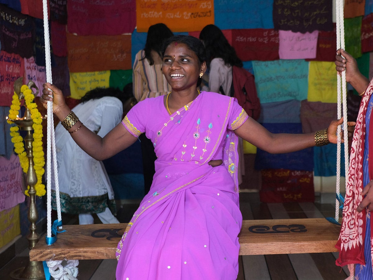 An Indian woman in a bright magenta sari smiles as she sits on a wooden swing in a small gallery space. On the walls are pieces of colorful square fabric with hand-stitched messages.